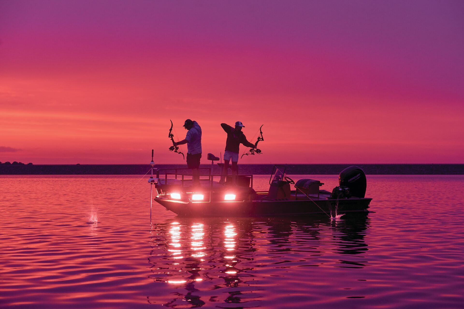 Two people on a boat at sunset, silhouetted against a pink and purple sky, holding bows for bowfishing.