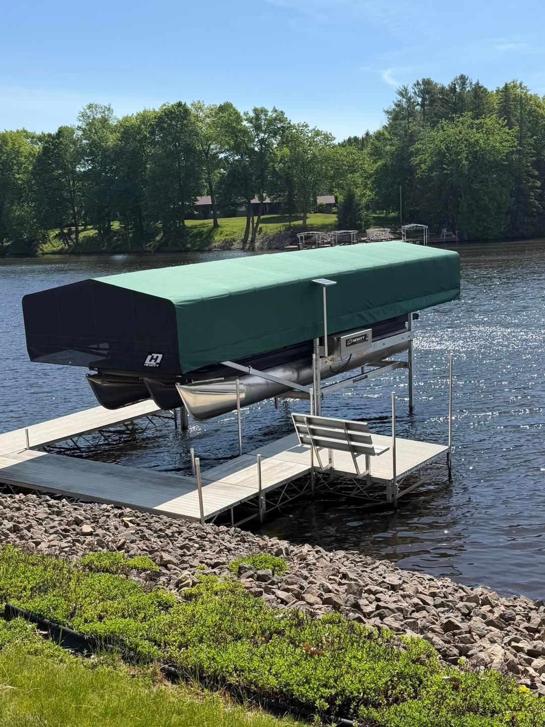 Covered boat lift with green canopy on a lake beside a grassy shoreline and trees.