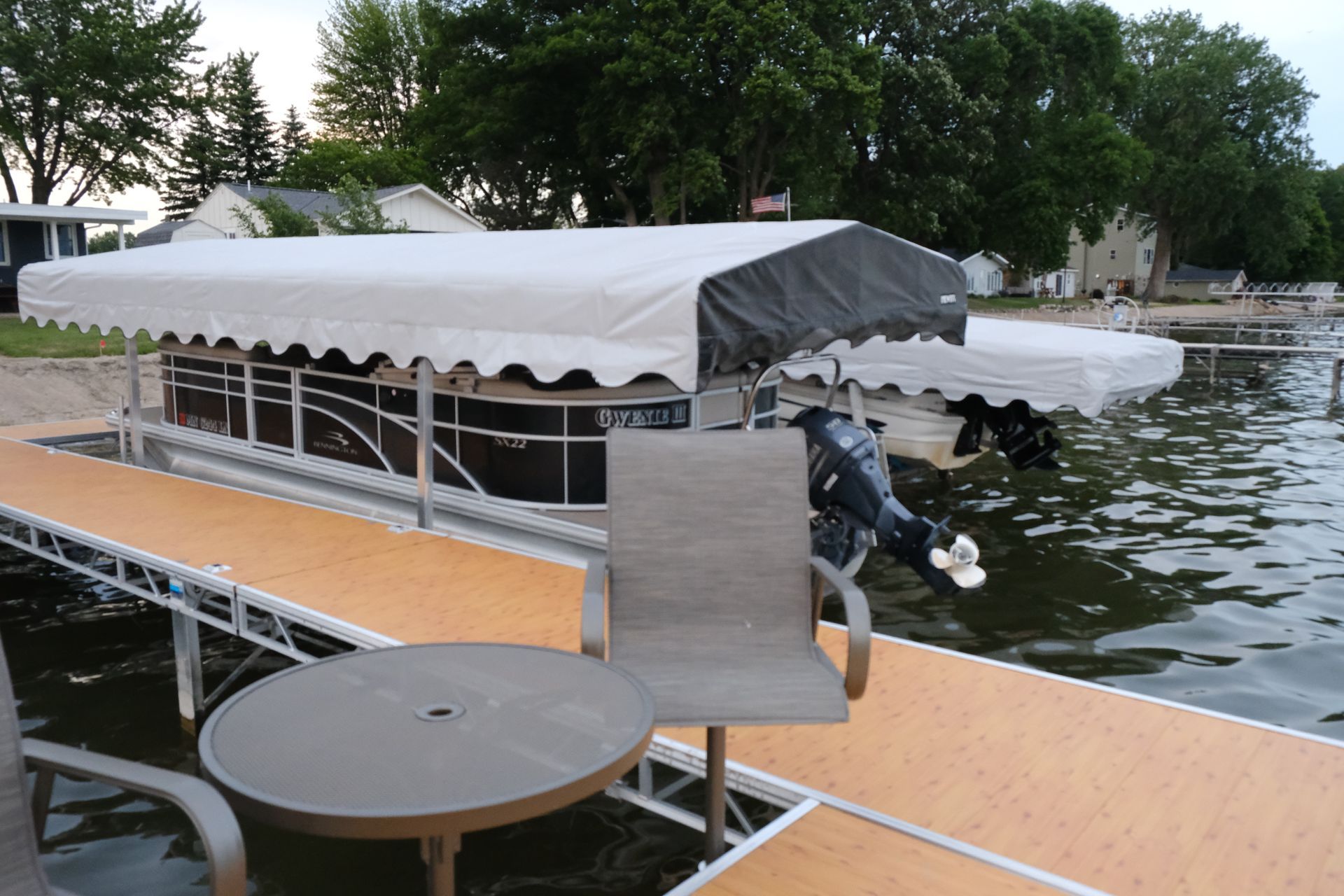 Dock with covered boat lift on a calm lakeside beside a round table and chairs, trees in the background.