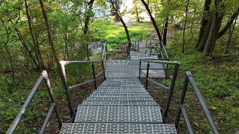 Steep metal stairway descending through a wooded forest with railings on both sides.
