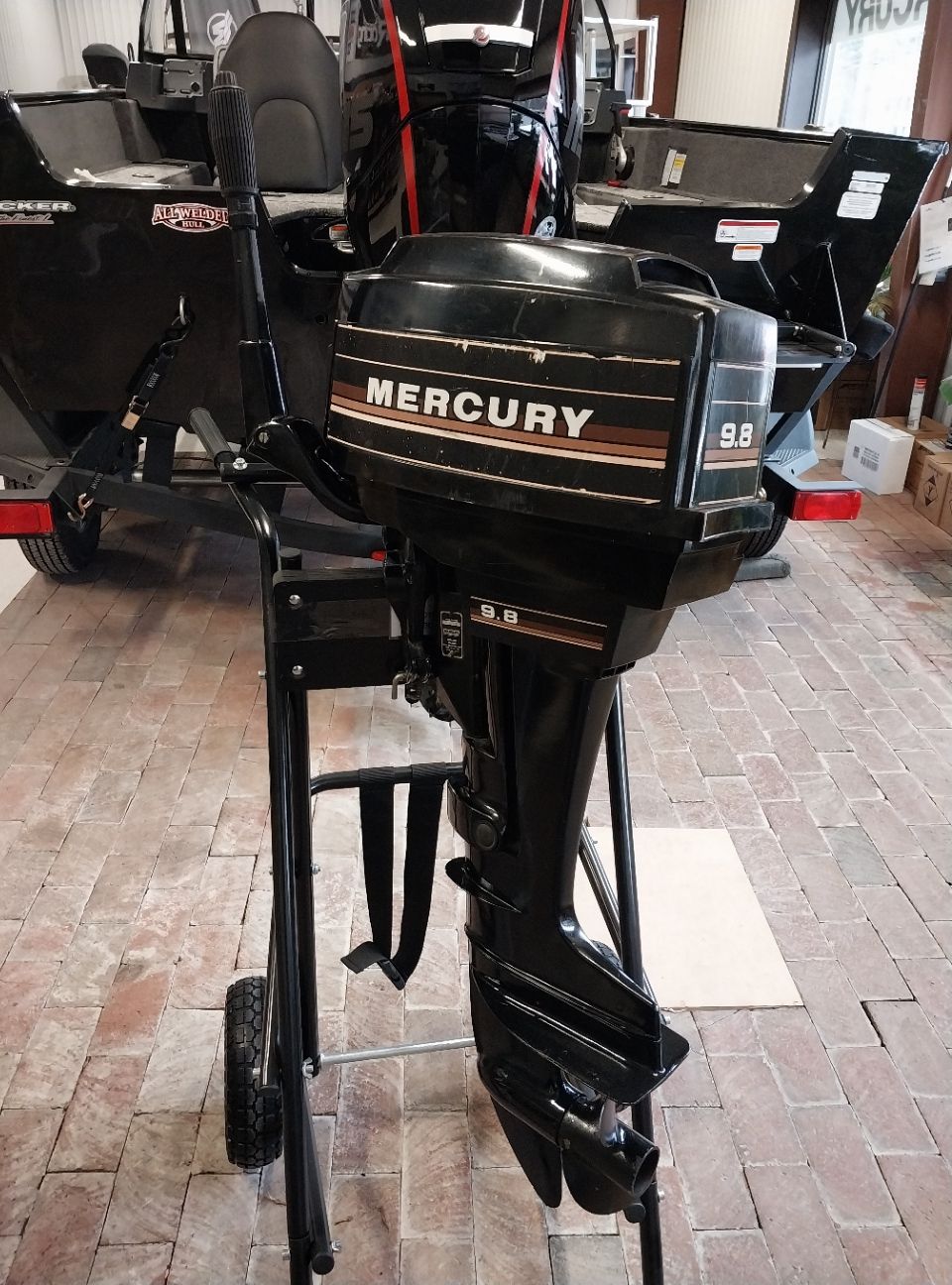 Black Mercury 9.8 outboard motor on a black metal stand, in front of a black boat on a trailer.