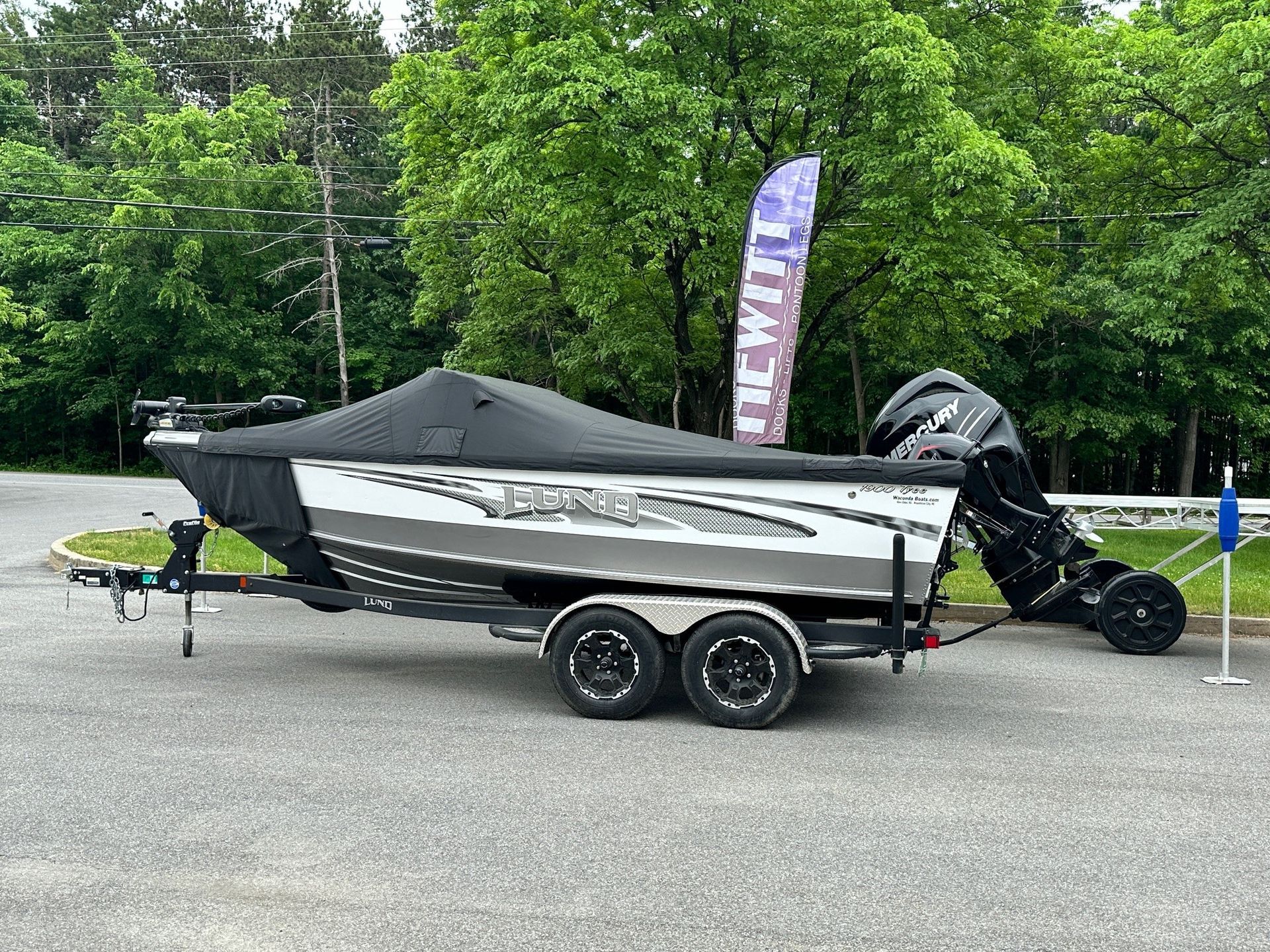 Boat on trailer, covered, parked on pavement; trees in background.