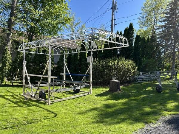 Metal boat lift frame on wheels, parked on green grass, next to trees and a road.