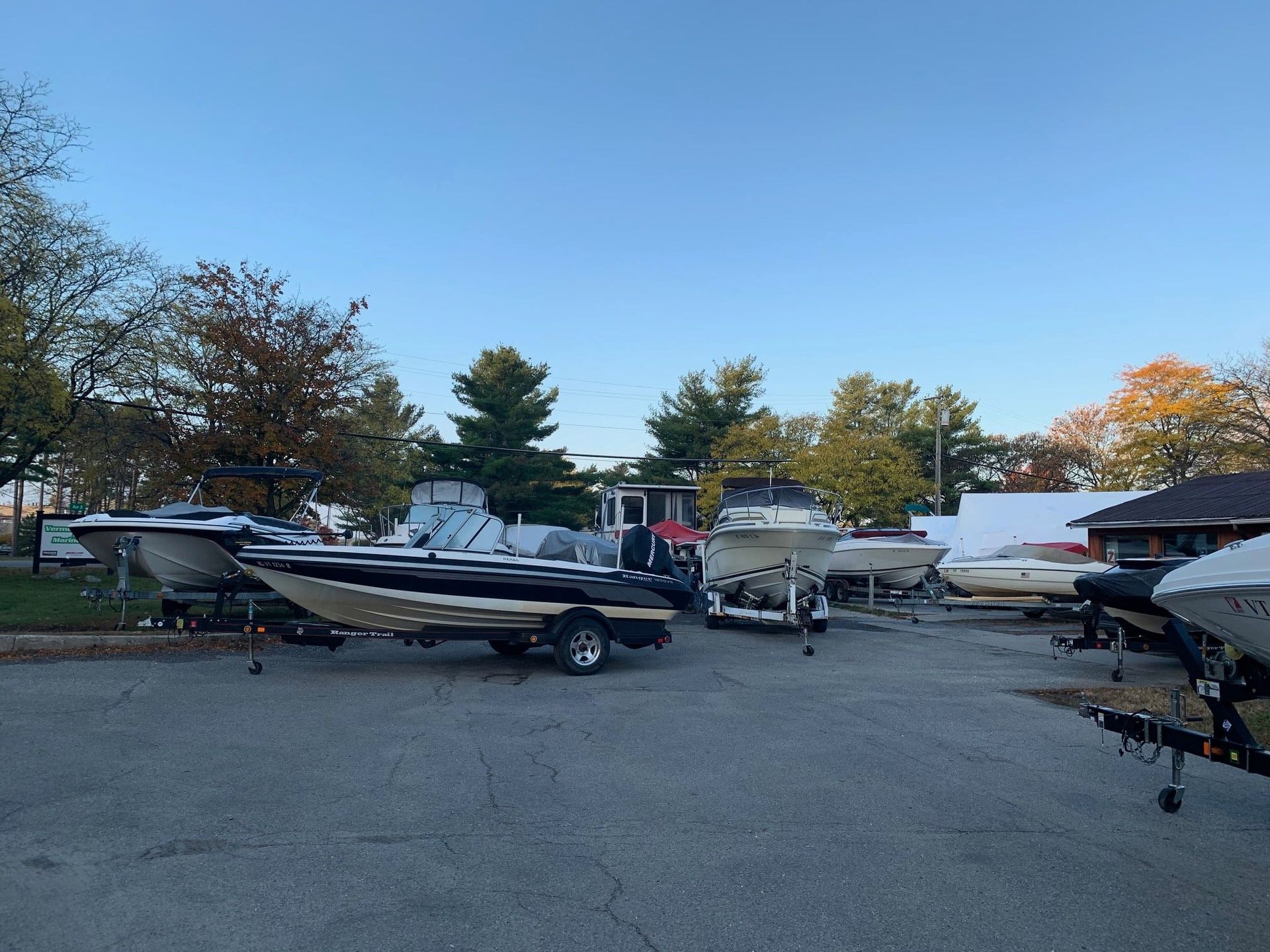 Boats on trailers in a paved lot under a blue sky, trees in the background.