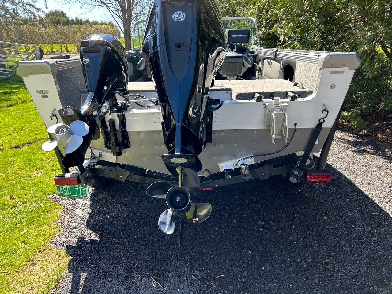 A boat with two black outboard motors, a green license plate, and silver hull on a driveway.