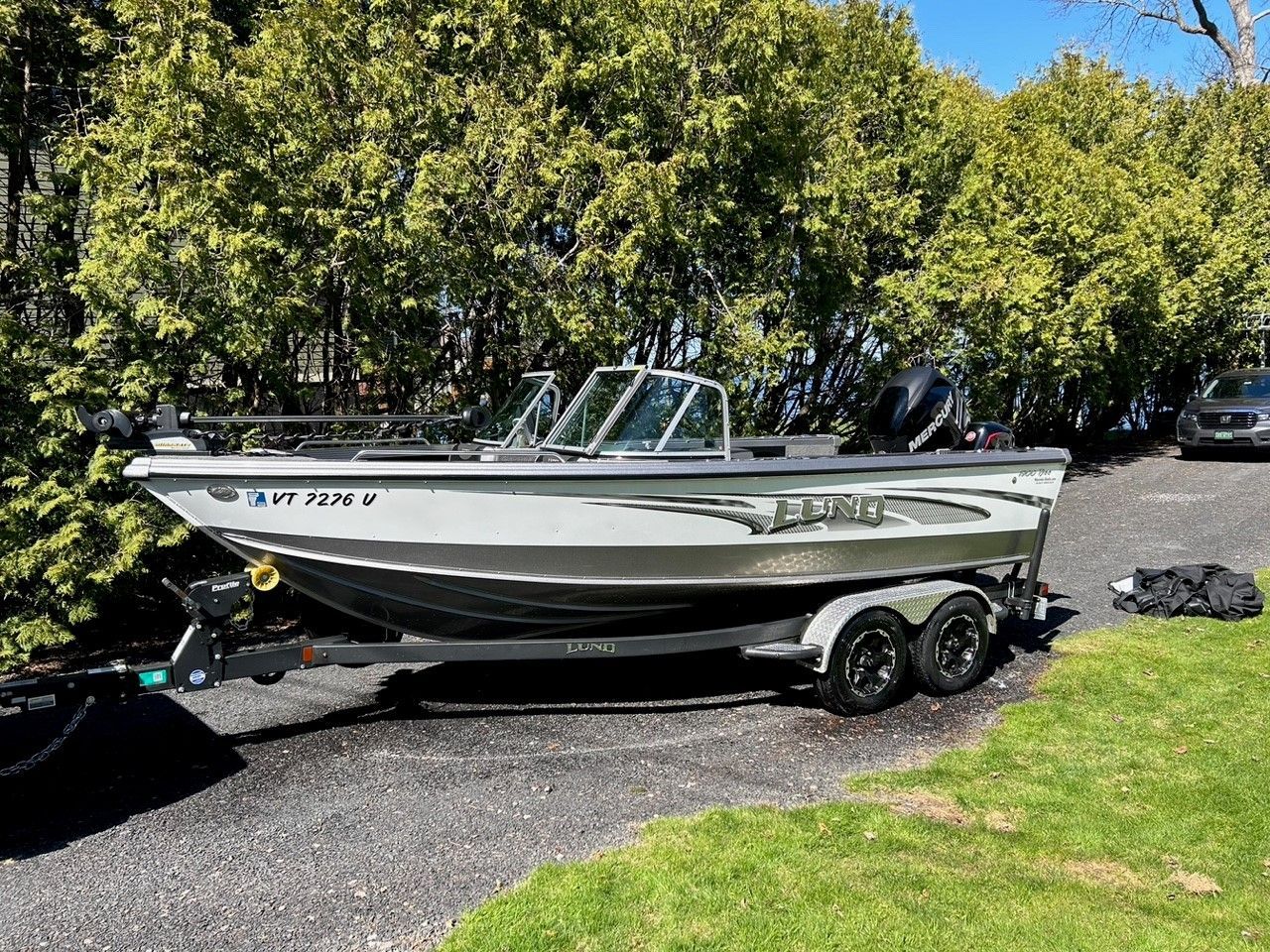 A silver and black fishing boat on a trailer parked on gravel, with green trees in the background.