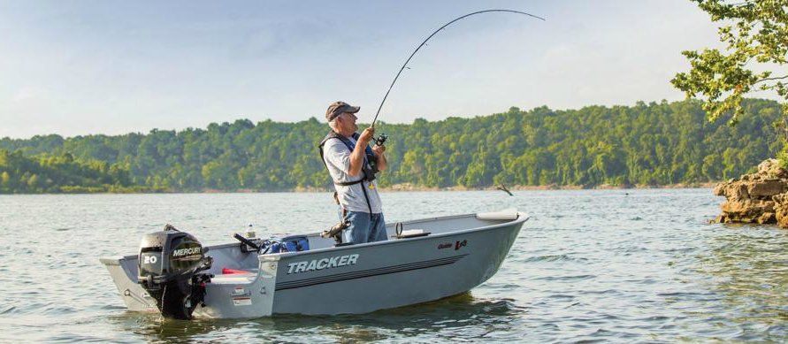 Man fishing from a small boat on a lake with green trees on the shore.