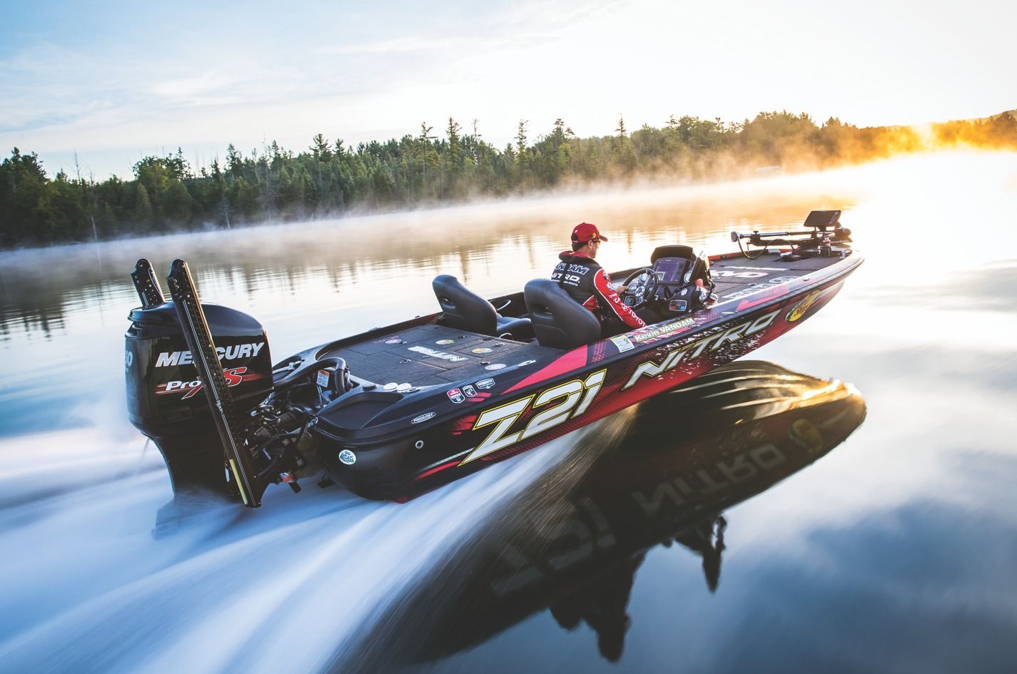 Bass boat speeding across water, driver in red cap, misty lake at sunrise.