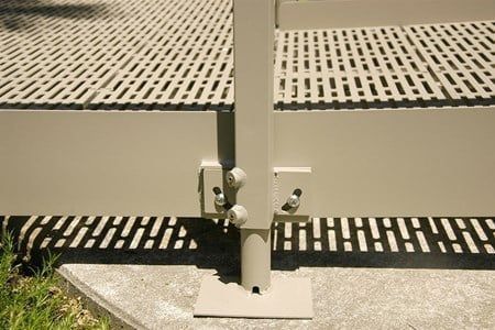 Beige metal platform with grid surface, bolted to a concrete base, casting shadows.