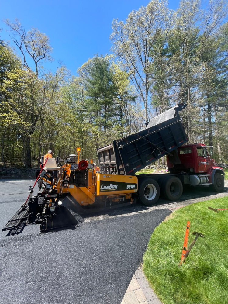A dump truck is being loaded with asphalt in a driveway.