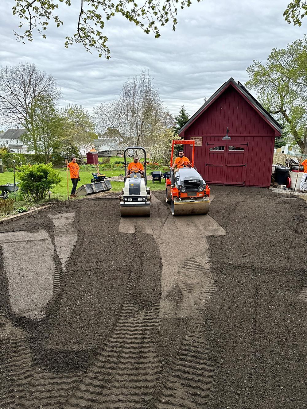 A group of people are working on a driveway in front of a red barn.