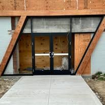 Exterior of a building with angled wood beams and black double doors. Concrete walkway in front.