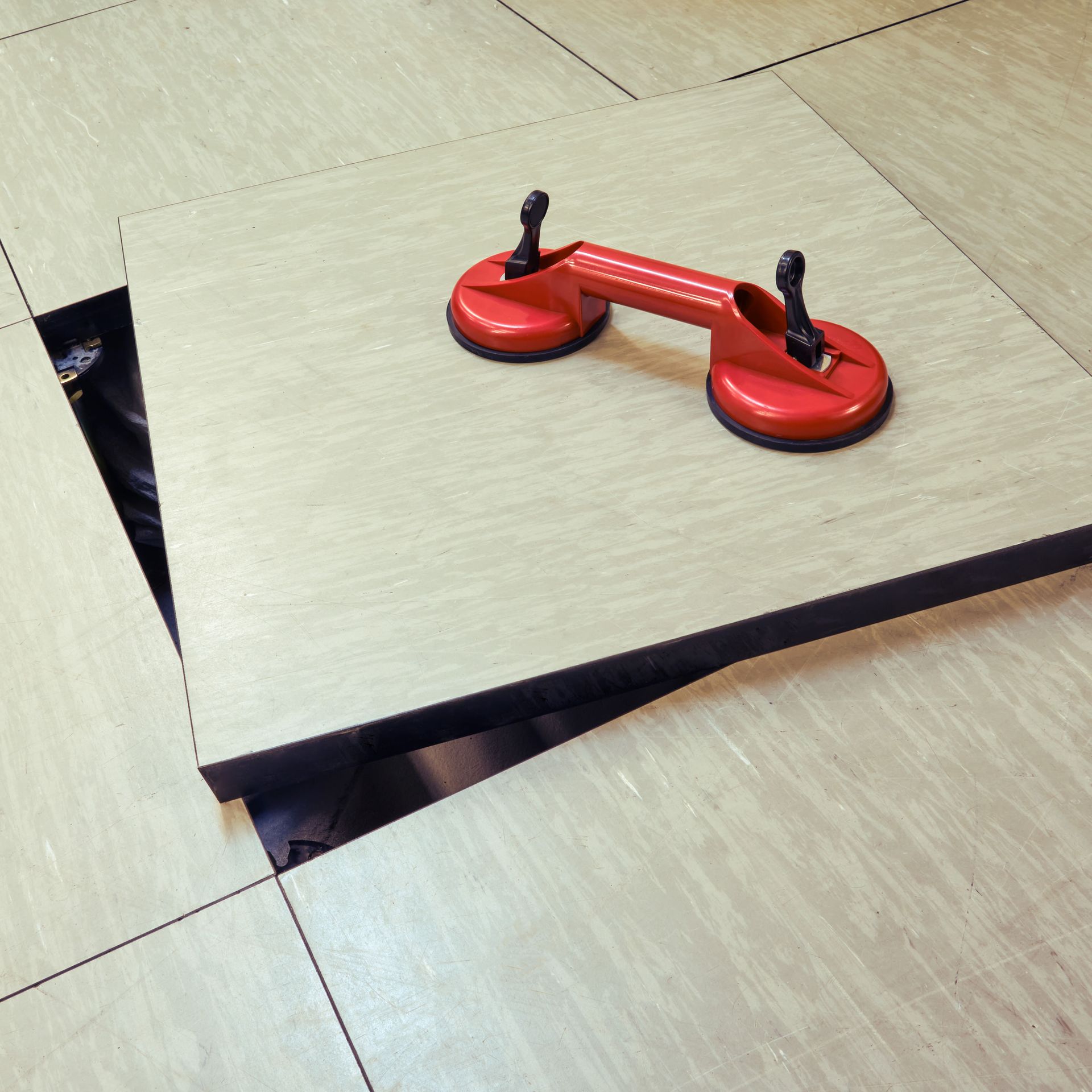 Raised floor panel being lifted with a red suction cup tool, revealing a dark access area.