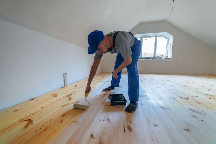 A person in blue overalls applying finish to a light wood floor in a room with a window.