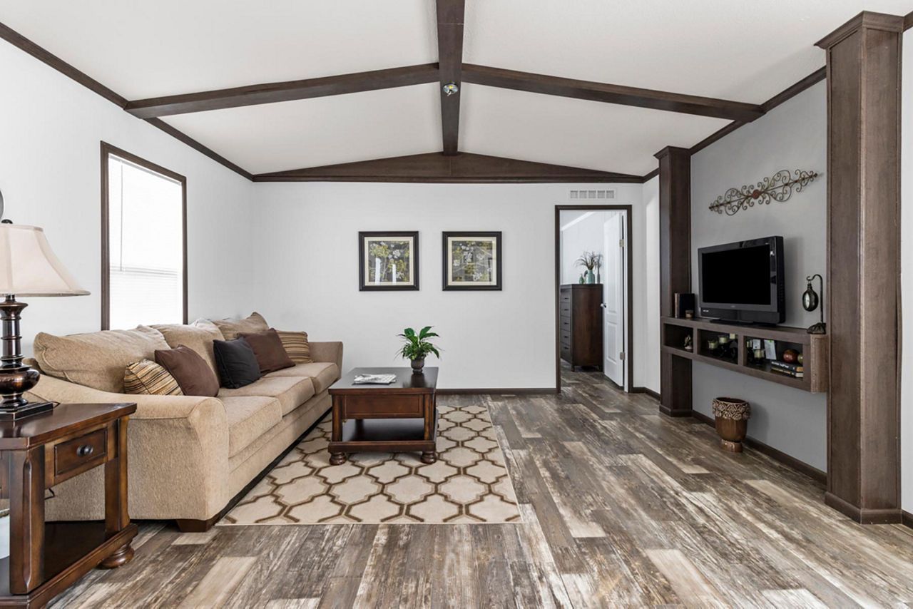 Living room interior with light-colored walls, dark wood beams, and a light brown sofa on a patterned rug.