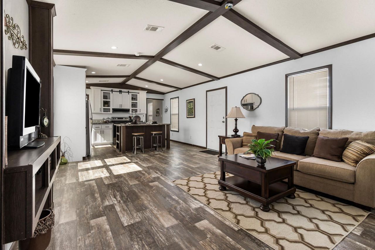 Spacious living room with dark wood flooring, a brown sofa, and a coffee table. A kitchen is visible in the background.