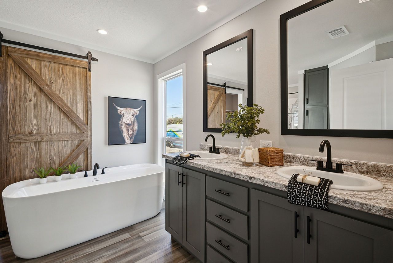 Bathroom with soaking tub, gray cabinets, two sinks, mirrors, and a wooden sliding barn door.