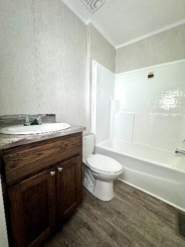 Bathroom with a wooden vanity, white toilet, and a white bathtub-shower combo. The walls are light gray and the floor is gray.