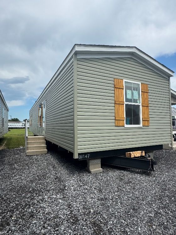 A light green mobile home with brown shutters, parked on gravel with a cloudy sky background.