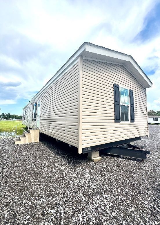 A beige manufactured home with black shutters sits on gravel under a cloudy sky.