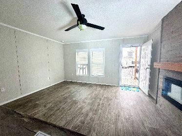 Empty living room with gray panel walls, wood-look flooring, and a ceiling fan. A door is open, leading outside.