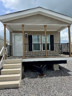 Tan-colored tiny home with a porch and steps.  Gray shutters, a white door, and a black railing are visible. It sits on a gravel surface.