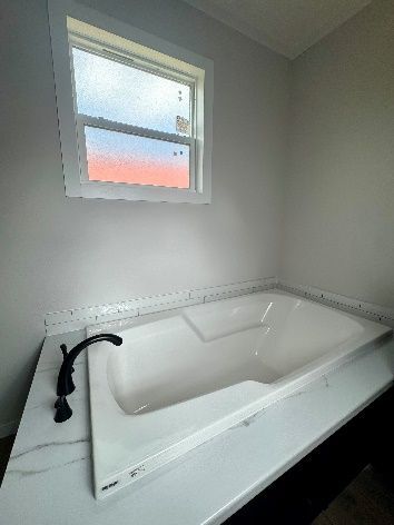 A white bathtub is built into a white countertop with black fixtures, next to a window in a bathroom.