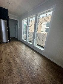 Room with wood-look flooring, three large windows with white frames, and a refrigerator in the background.