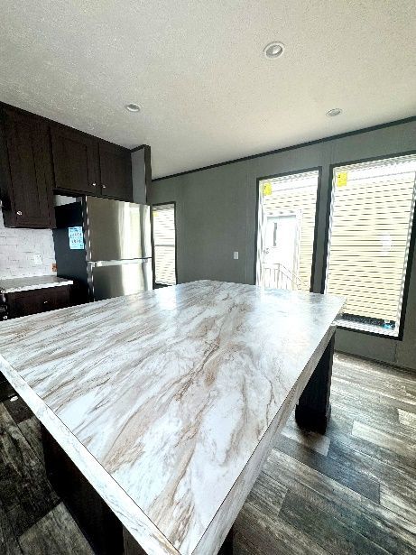 Interior view of a modern kitchen featuring a large island with a marble-look countertop. Dark cabinets and a stainless steel refrigerator are visible.