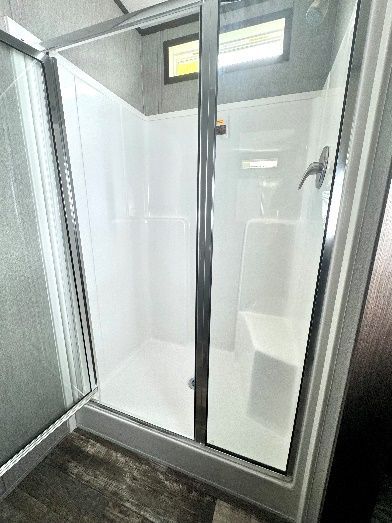 A shower with a glass door, white walls, and a built-in seat inside. The floor has dark wood-look tiles.