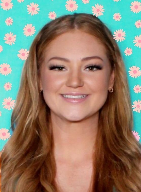 A woman with long red hair is smiling in front of a blue background with pink daisies