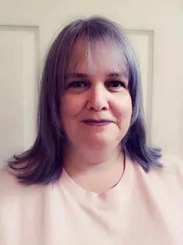 A woman with purple hair is smiling and wearing a pink shirt.
