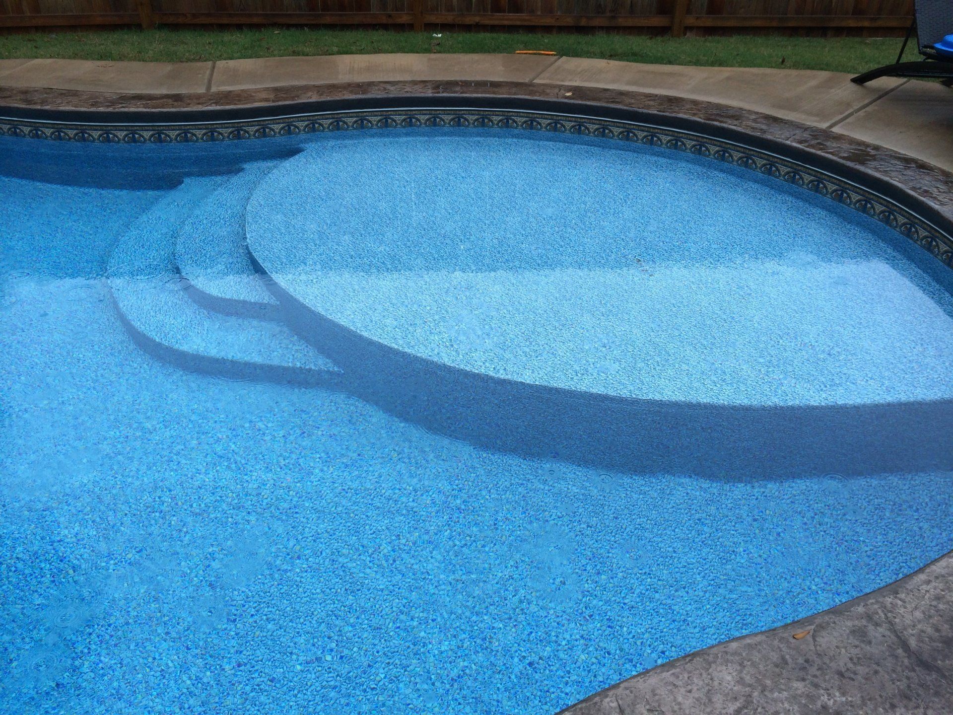 A blue swimming pool with stairs and a fence in the background.