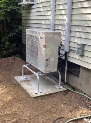 Air conditioner unit on concrete pad next to a light-colored house. Gray support structure and piping.
