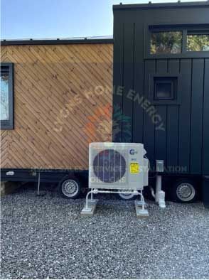 Air conditioning unit installed next to a tiny home with black and wood siding on a gravel surface.