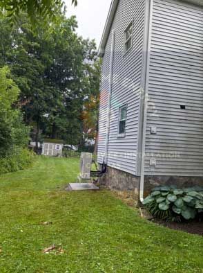 Side view of a light gray house with a stone foundation and green grass yard, trees in the background.