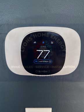 A digital thermostat displaying 77 degrees, mounted on a blue wall, with Lyons Home Energy branding.