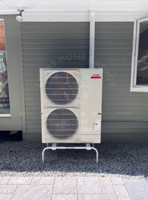 Mitsubishi heat pump mounted outside on gravel, next to a building with a white pipe.