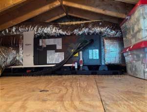 HVAC unit in an attic with ductwork, set on plywood flooring. Storage bins on the right.
