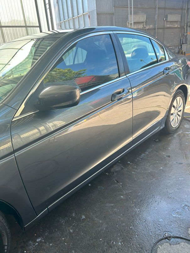 Dark gray sedan parked on a wet driveway beside a building, with sunlight reflecting on the doors.