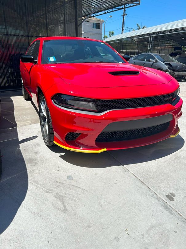 Red sports car parked outdoors in bright sunlight, front view with black grille and hood scoop.