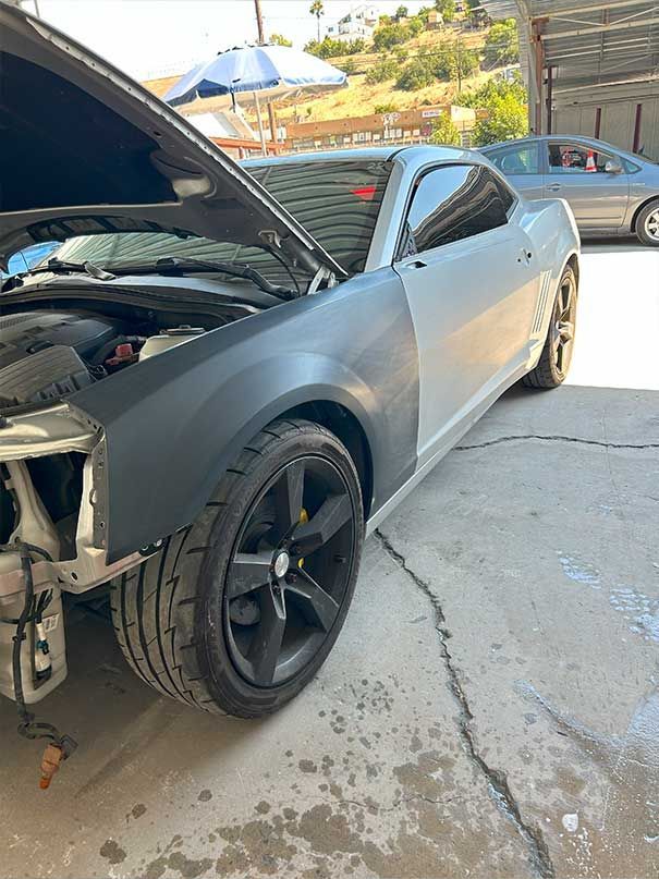 Damaged gray sports car with open hood and missing front bumper, parked on a driveway.