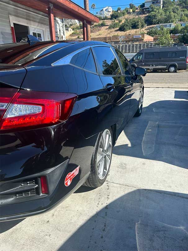 Black sedan parked in a sunny driveway beside a house, with a van in the background.