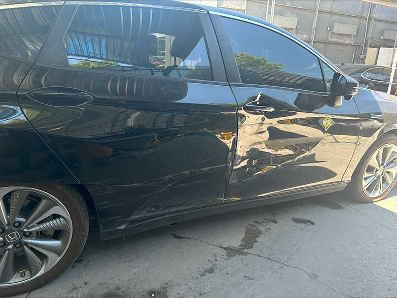 Black sedan with side damage parked in bright sunlight, showing dented doors and a damaged front wheel area