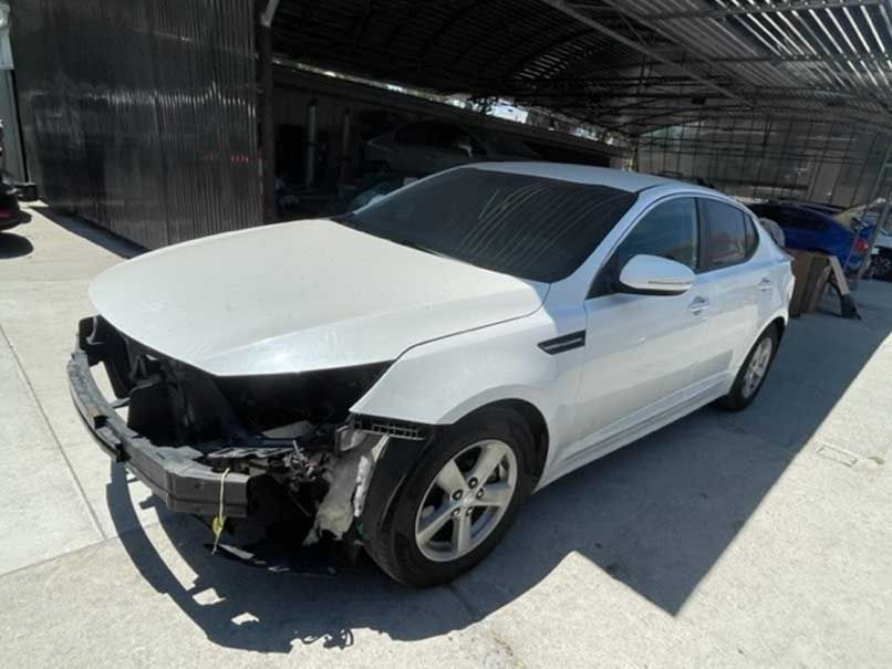 White sedan with front-end damage, missing bumper, parked outdoors in bright sunlight