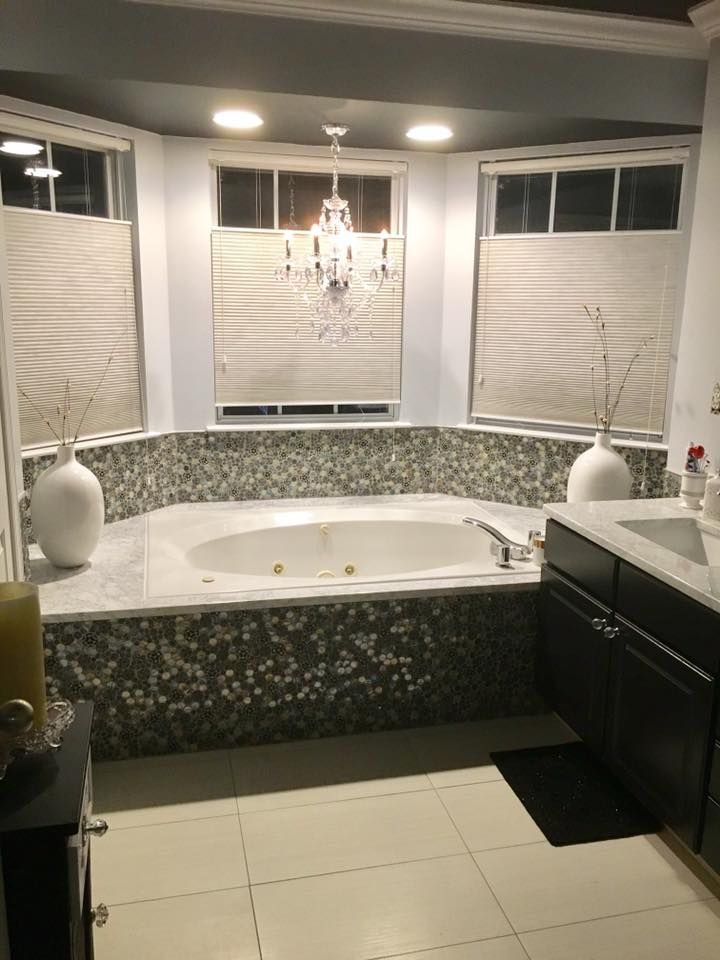 Luxury bathroom with a large corner bathtub, chandelier, mosaic tile surround, and white blinds on the windows