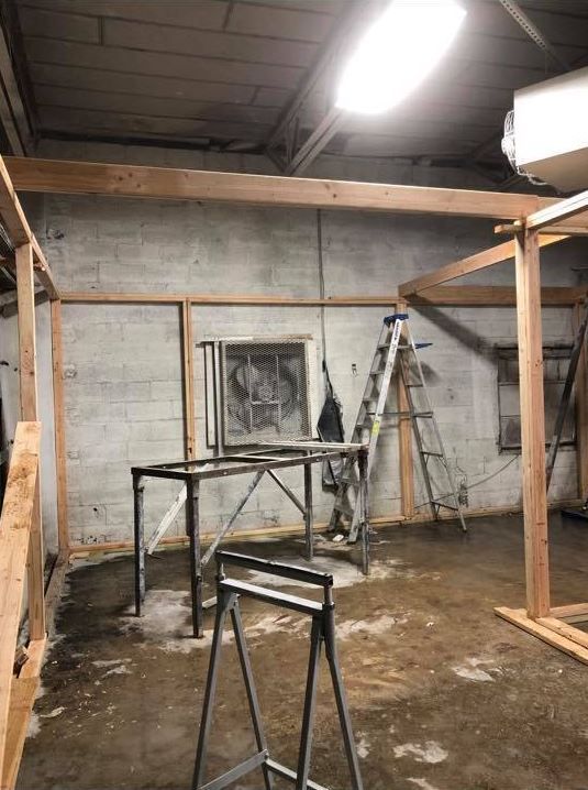 Dim workshop with cinderblock walls, wooden framing, metal sawhorses, ladder, and overhead light.
