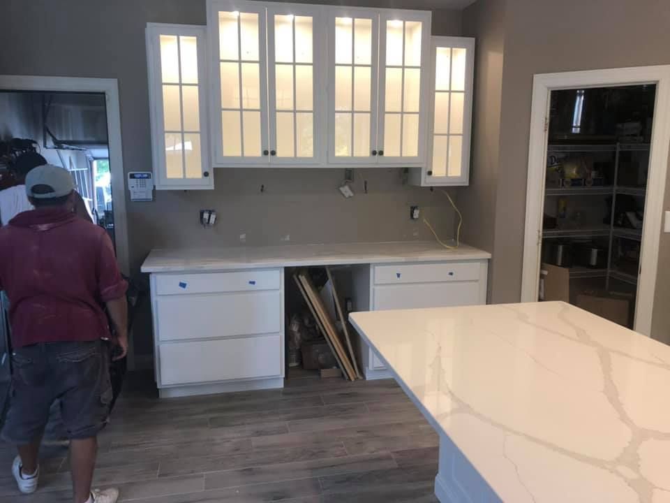 Kitchen with white cabinets and marble island; person in maroon shirt stands at left near an open pantry.