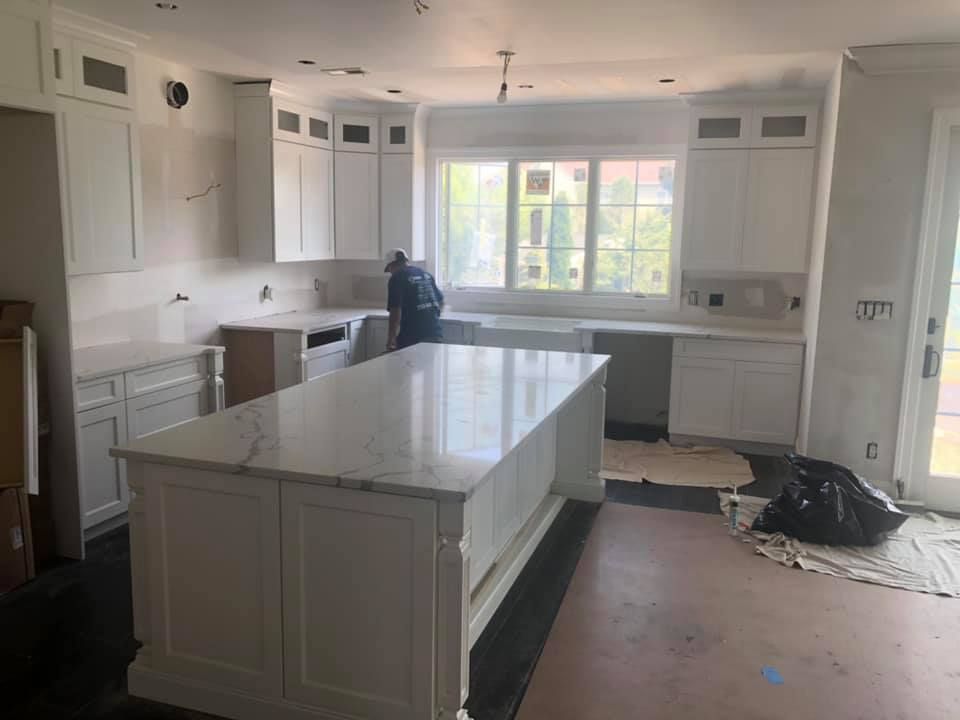 Kitchen under renovation with white cabinets, large island, and unfinished floor near a bright window.
