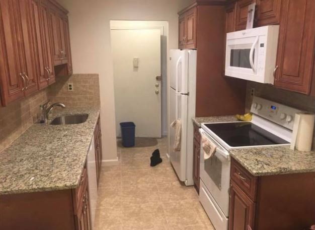 Narrow kitchen with wood cabinets, granite counters, white appliances, and a blue bin by the back door
